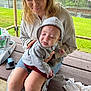 baby, woman, outdoor, bench, hoodie, grass, fence, basketball_court, casual, smile, tongue_out, jeans, ripped_jeans, holding, person, child, expression, daylight, wooden_bench, clothing