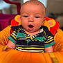 baby, child, infant, face, expression, striped_shirt, polo_shirt, orange_cushion, seat, indoors, blurred_background, wide_eyes, open_mouth, short_hair, cute, sitting, colorful_clothing, young_child, portrait, soft_lighting