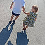 children, holding_hands, street, shadows, sunlight, boy, girl, white_shirt, shorts, dress, sunflower_pattern, walking, asphalt, sidewalk, urban, daytime, curved_road, cars, shoes, outdoor