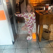 Arabella is registered to the contest to win money with this photo: child, girl, pajamas, slippers, heart_pattern, smiling, kitchen, stove, tile_floor, wooden_table, basket, indoor, happy, posing, cozy, home, appliance, cup, glass, countertop