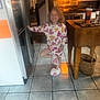 child, girl, pajamas, slippers, heart_pattern, smiling, kitchen, stove, tile_floor, wooden_table, basket, indoor, happy, posing, cozy, home, appliance, cup, glass, countertop