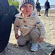 Jasper joined the competition — help win amazing prizes! toddler, child, beanie, hat, jacket, sweatpants, shoes, dinosaur_print, sand, beach, outdoor, daylight, water, people, background, nature, trees, sky, curious, crouching