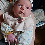 Mya a rejoint le concours — aidez-le/la à gagner de superbes lots ! baby, infant, newborn, headband, bib, blue_eyes, drool, hand, arm, lace_cuff, onesie, pattern, person, portrait, indoor, caregiver, blanket, bokeh_background, cozy, closeup