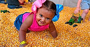 Marcella is registered to the contest to win money with this photo: child, children, toddler, girl, smiling, crawling, portrait, outdoor, corn, corn_kernels, play_area, toy_tractor, toys, hair_bows, pink_clothing, blue_clothing, playground, shallow_depth_of_field, happy, festival