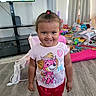 child, toddler, girl, smiling, portrait, standing, indoors, playroom, toys, television, curtains, floor, t_shirt, shorts, pink_clothing, hair_bow, curly_hair, furniture, home, happy