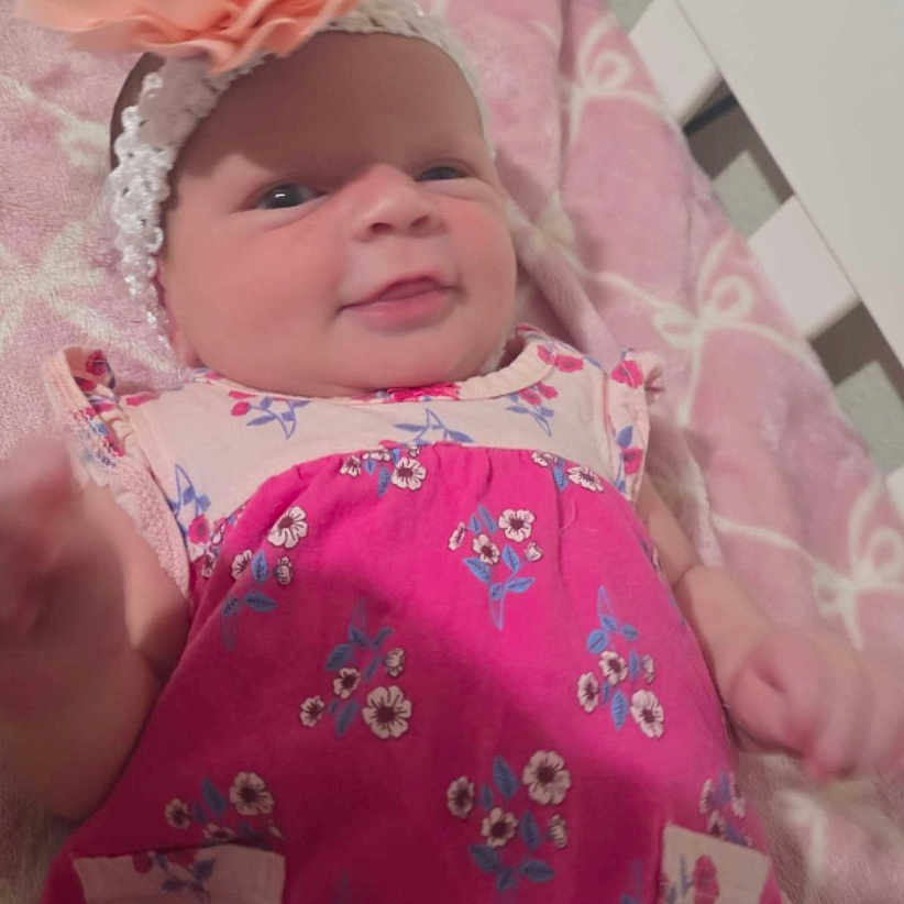 Chloe is registered to the contest to win money with this photo: baby, infant, crib, blanket, headband, flower, pink_clothing, floral_pattern, smile, face, child, indoors, furniture, bed, cute, newborn, lying_down, person, soft_toys, cozy