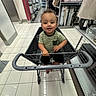 toddler, shopping_cart, store, aisle, floor_tiles, shelves, bedding, lamp, curious, child, indoor, clothing, striped_shirt, black_shorts, person, smile, shopping, retail, baby, handle