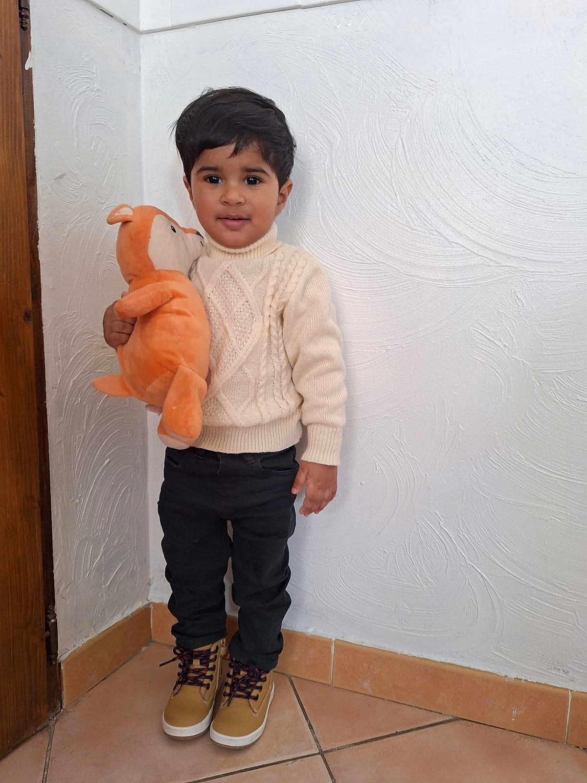 Ilyan participe au concours pour gagner de l'argent avec cette photo : child, toddler, boy, plush_toy, stuffed_animal, sweater, turtleneck_sweater, black_pants, boots, standing, indoor, textured_wall, tile_floor, doorway, portrait, smile, brown_hair, hand, cute, cozy