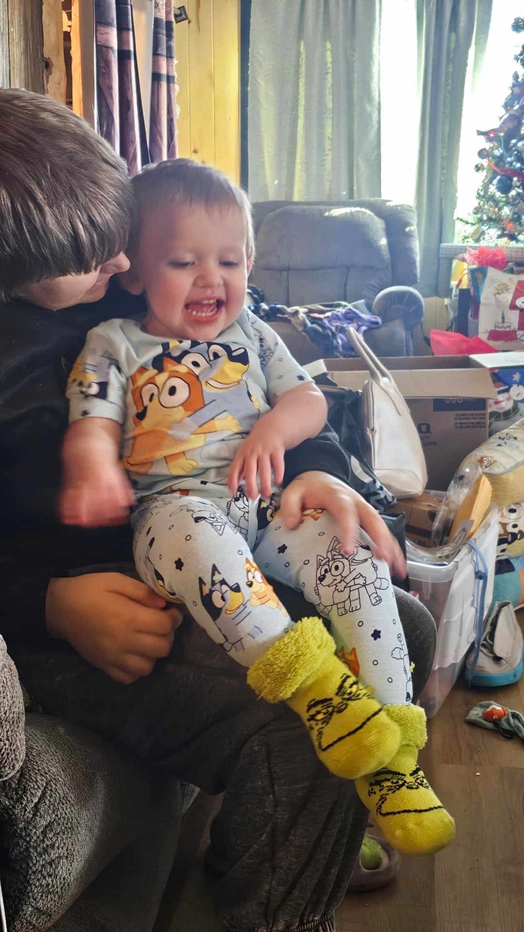 Kameron is registered to the contest to win money with this photo: toddler, child, pajamas, yellow_socks, smiling, laughing, indoor, living_room, sofa, curtains, holiday_decor, christmas_tree, boxes, hand, person, clothing, carpet, furniture, family, cozy