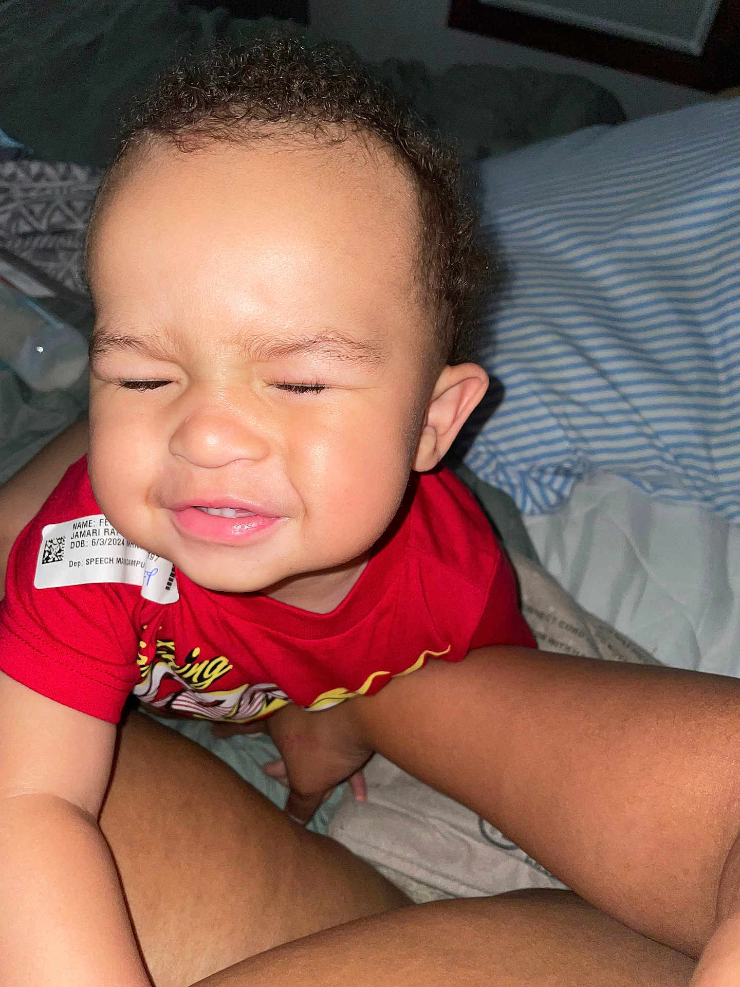 Jenesis is registered to the contest to win money with this photo: toddler, child, face, smile, red_shirt, tag, curly_hair, bed, pillow, striped_pillow, legs, person, indoor, closeup, expression, cute, skin, baby, cheek, hand