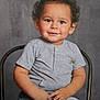 child, toddler, smiling, curly_hair, grey_shirt, jeans, sitting, chair, portrait, casual_clothing, happy, cute, indoor, studio, young_child, person, face, expression, background, hands