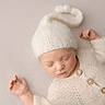 baby, sleeping, newborn, knit_hat, knit_sweater, buttons, soft_texture, cozy, neutral_background, peaceful, infant, portrait, cute, child, hands, closed_eyes, warm, resting, delicate, gentle_lighting