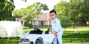 Kendrys is registered to the contest to win money with this photo: toddler, child, toy_car, white_suit, outdoor, park, fountain, greenery, sidewalk, miniature_car, luxury_toy, confident, portrait, formal_wear, summer, daylight, young_child, black_shoes, nature, cute