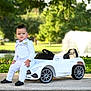 toddler, child, toy_car, white_suit, outdoor, park, fountain, greenery, sidewalk, black_shoes, person, portrait, sunlight, nature, grass, trees, casual_pose, young_child, fashion, playtime