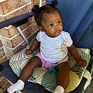 Amoura joined the competition — help win amazing prizes! baby, child, toddler, sitting, chair, folding_chair, cushion, pacifier, white_shirt, pink_shorts, socks, curly_hair, pigtails, portrait, outdoor, brick_wall, patio, infant, looking_away, patterned_fabric