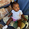 baby, child, toddler, sitting, chair, folding_chair, cushion, pacifier, white_shirt, pink_shorts, socks, curly_hair, pigtails, portrait, outdoor, brick_wall, patio, infant, looking_away, patterned_fabric