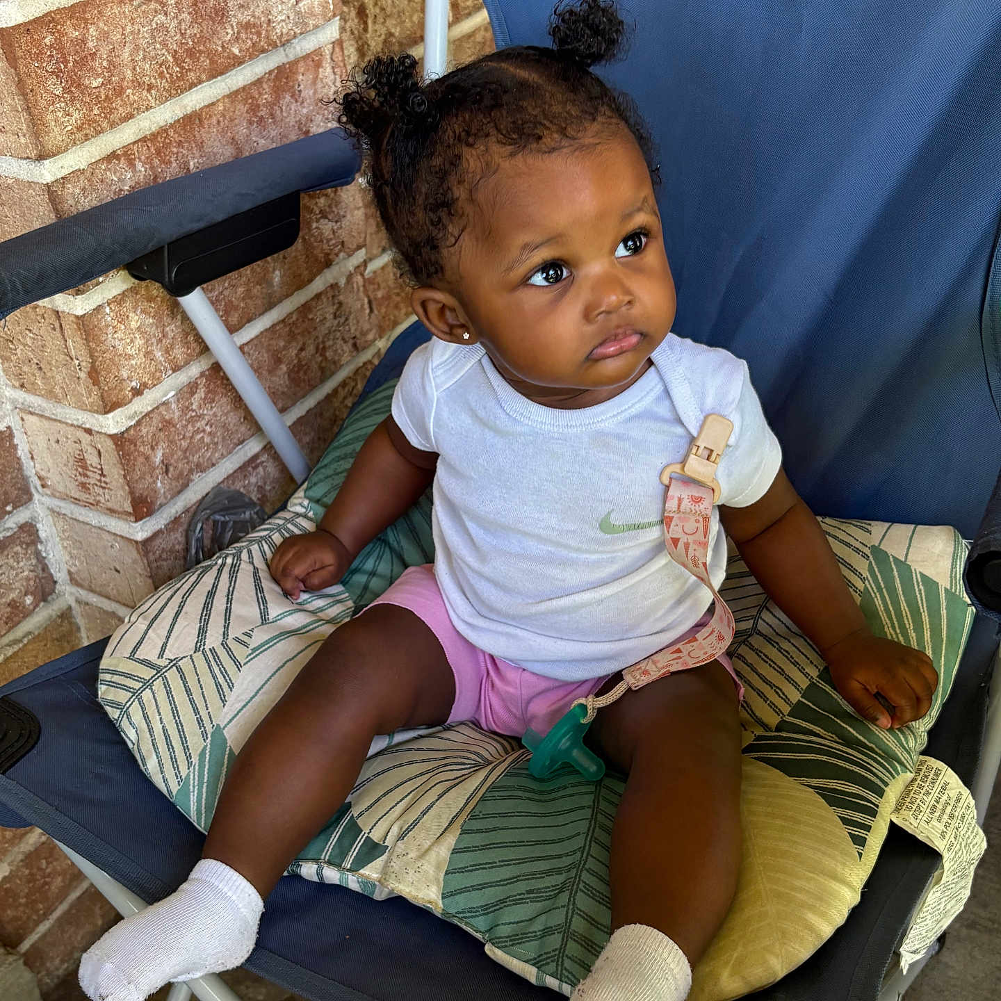 Amoura joined the competition — help win amazing prizes! baby, child, toddler, sitting, chair, folding_chair, cushion, pacifier, white_shirt, pink_shorts, socks, curly_hair, pigtails, portrait, outdoor, brick_wall, patio, infant, looking_away, patterned_fabric