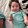 baby, child, smiling, curly_hair, onesie, bed, blanket, happy, person, infant, cute, portrait, lying_down, indoor, clothing, face, skin, hand, blue, text_on_clothing