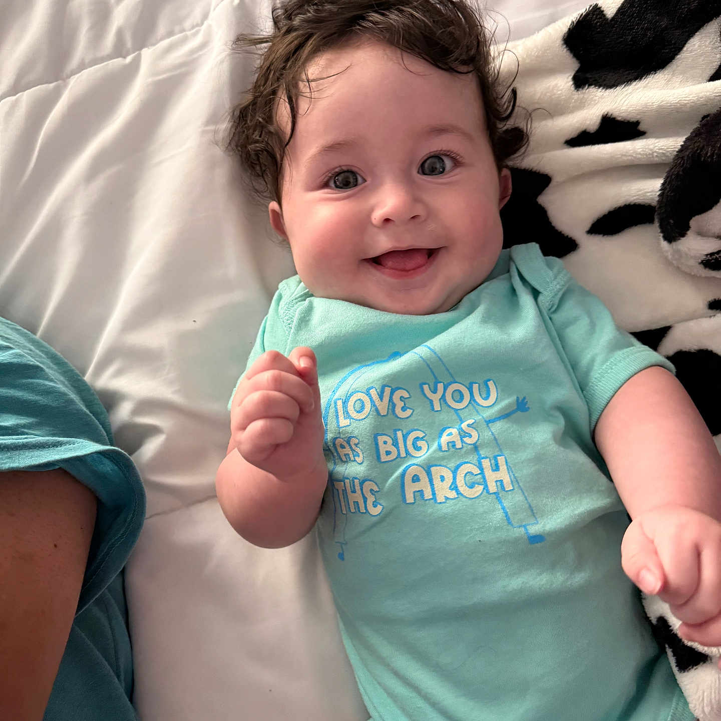 Nova is registered to the contest to win money with this photo: baby, child, smiling, curly_hair, onesie, bed, blanket, happy, person, infant, cute, portrait, lying_down, indoor, clothing, face, skin, hand, blue, text_on_clothing
