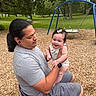 adult, baby, playground, swing, park, grass, trees, child, smile, happy, outdoor, nature, casual_clothing, hair, person, seat, woodchips, accessory, bracelet, watch