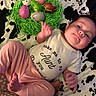 baby, child, infant, onesie, pink_pants, plastic_eggs, green_shredded_paper, cow_print_blanket, face, smile, hand, foot, indoor, cute, playful, easter, laying_down, clothing, person, portrait