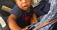 Trillion is registered to the contest to win money with this photo: toddler, child, baby, stroller, indoor, person, curly_hair, blue_shirt, expression, wide_eyes, hand, face, seat, furniture, adult_in_background, door, floor, warning_label, casual_clothing, portrait