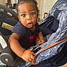 toddler, child, baby, stroller, indoor, person, curly_hair, blue_shirt, expression, wide_eyes, hand, face, seat, furniture, adult_in_background, door, floor, warning_label, casual_clothing, portrait