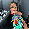 Trillion joined the competition — help win amazing prizes! toddler, child, car_seat, toy, bib, smiling, black_hair, curly_hair, black_clothing, happy, indoors, seatbelt, car_interior, playful, cute, baby, person, sitting, bright_colors, portrait