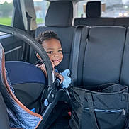 Trillion is registered to the contest to win money with this photo: toddler, child, car_seat, vehicle_interior, smile, face, person, seat, black_bag, window, curly_hair, car, seatbelt, daylight, backseat, happy, indoor, transportation, baby, holding