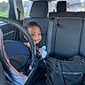 toddler, child, car_seat, vehicle_interior, smile, face, person, seat, black_bag, window, curly_hair, car, seatbelt, daylight, backseat, happy, indoor, transportation, baby, holding