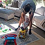 Bentley is registered to the contest to win money with this photo: baby, adult, toy_bus, living_room, carpet, sofa, cushion, window, plant, rug, floor, toy, child, person, playtime, sunlight, home_interior, support, smile, casual_clothing