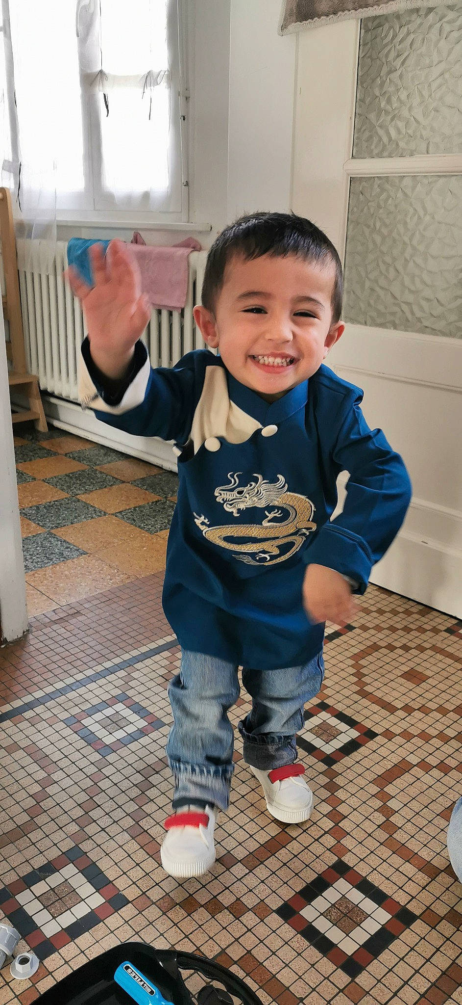 May participe au concours pour gagner de l'argent avec cette photo : baby_toddler_clothing, child, electric_blue, floor, flooring, fun, gesture, happy, hardwood, head, human_leg, joy, person, play, sitting, skin, sleeve, smile, t_shirt, thumb