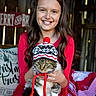 Squish is registered to the contest to win money with this photo: blanket, cat, christmas_decor, cozy, ear_flaps, festive, furry_pet, girl, hat, holding, holiday, indoor, pet, pillow, pom_pom, portrait, red_dress, smiling, winter_hat, wooden_wall