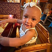 Summer is registered to the contest to win money with this photo: toddler, child, high_chair, restaurant, table, wooden_furniture, headband, bow, smile, curious, indoor, warm_lighting, face, arm, clothing, person, baby, seat, happy, cute
