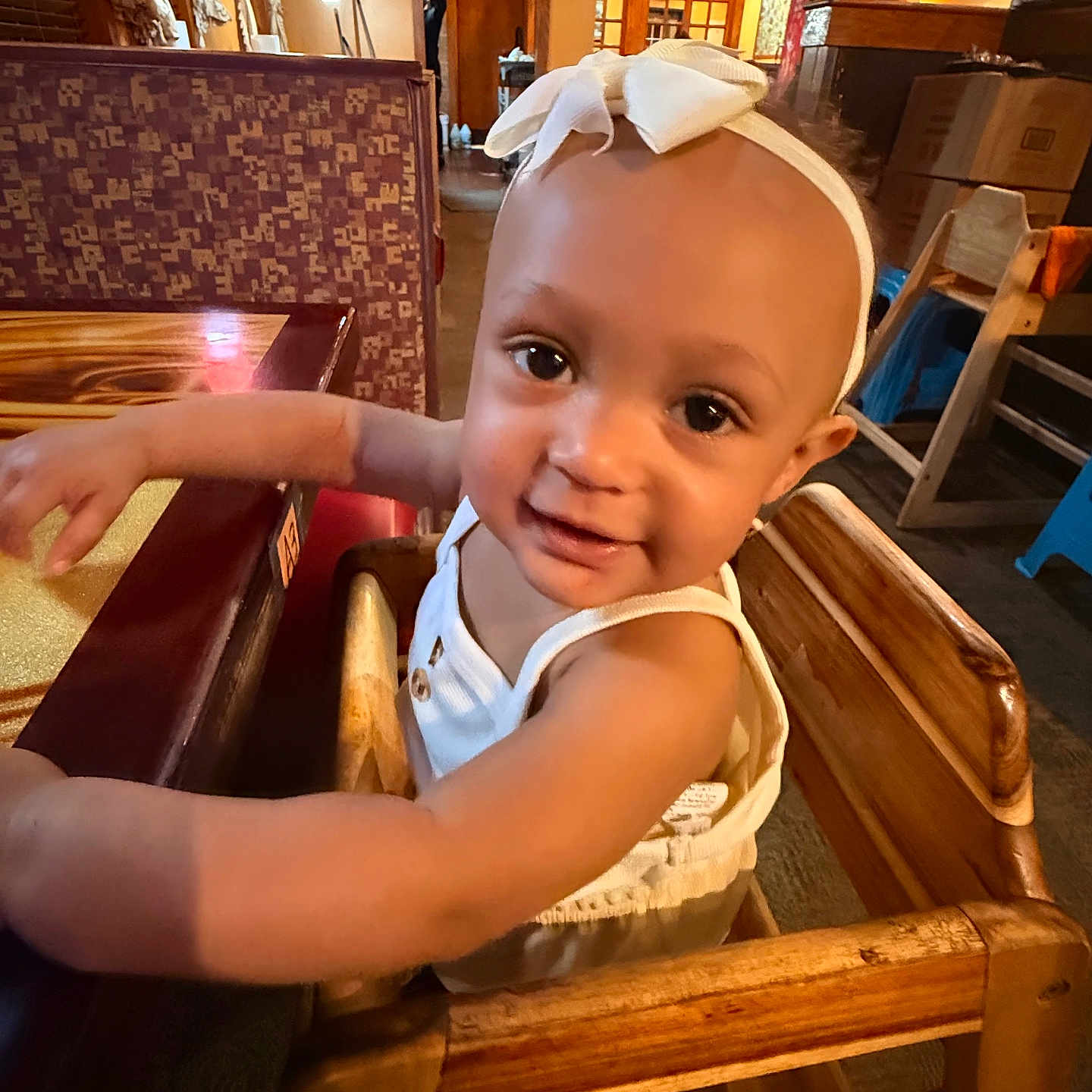 Summer is registered to the contest to win money with this photo: toddler, child, high_chair, restaurant, table, wooden_furniture, headband, bow, smile, curious, indoor, warm_lighting, face, arm, clothing, person, baby, seat, happy, cute
