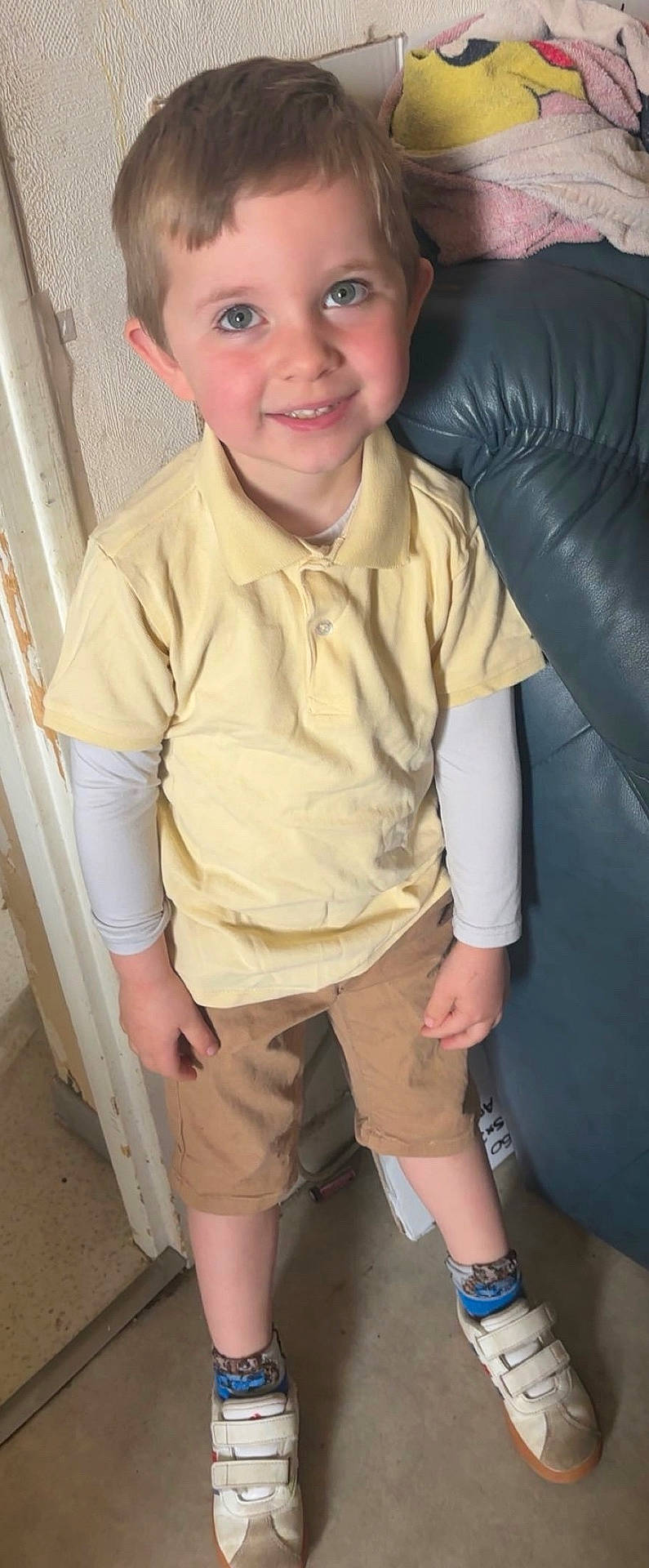 blanket, blue_couch, boy, casual_clothing, child, doorway, floor, happy, home, indoor, long_sleeve_shirt, portrait, shorts, smiling, sneakers, socks, standing, wall, yellow_shirt, young_child