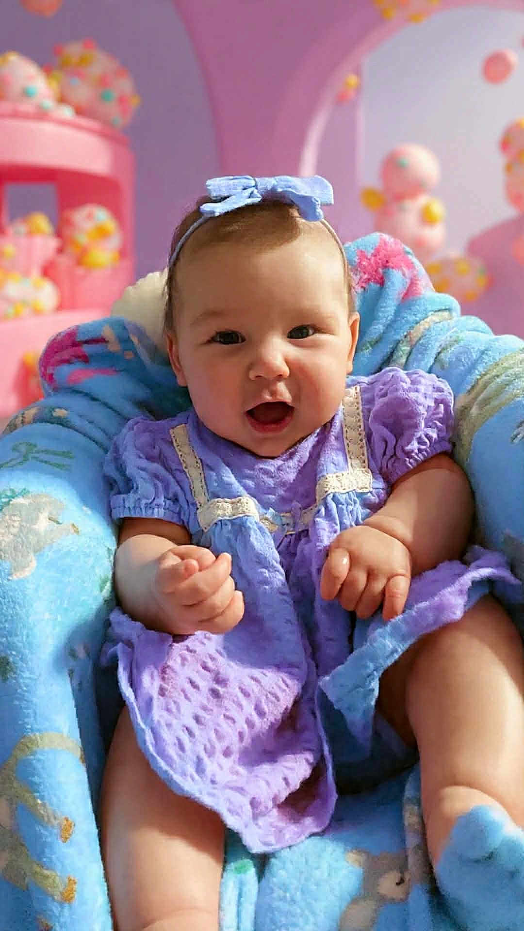 Kylie is registered to the contest to win money with this photo: baby, child, infant, purple_dress, headband, blanket, blue_blanket, smiling, happy, indoor, soft_toys, pastel_colors, pink_background, cute, chubby_arms, seated, portrait, cozy, warm, adorable
