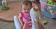 Nova is registered to the contest to win money with this photo: toddler, child, rocking_toy, swan, plush, indoor, floor, toy_box, colorful_toys, pink_clothing, furniture, wooden_floor, happy, playtime, smiling, baby, head, face, legs, room