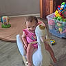 toddler, child, rocking_toy, swan, plush, indoor, floor, toy_box, colorful_toys, pink_clothing, furniture, wooden_floor, happy, playtime, smiling, baby, head, face, legs, room