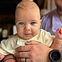 baby, child, blue_eyes, close_up, holding, adult_hand, watch, ring, portrait, indoor, soft_lighting, cute, infant, focus, person, skin, face, expression, background_blur, casual