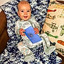 baby, child, blue_blanket, brown_chair, colorful_pillow, decorative_pillow, face, hand, infant, indoors, knit_blanket, onesie, patterned_clothing, person, plush, sitting, smiling, toy, white, blue
