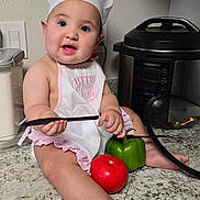 Adriana is registered to the contest to win money with this photo: baby, child, chef_hat, apron, kitchen, countertop, knife, green_bell_pepper, tomato, pressure_cooker, cooking, cute, infant, playing, food, person, indoor, curious, sitting, smiling