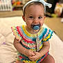 baby, child, pacifier, headband, crochet_dress, colorful_clothing, sitting, indoor, smiling, cute, hands_clasped, infant, portrait, young_child, soft_focus, happy, person, baby_girl, playroom, blanket