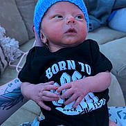 Ryder is registered to the contest to win money with this photo: newborn, baby, blue_hat, black_onesie, tattoo, couch, indoor, person, infant, hand, face, clothing, sitting, cute, portrait, skin, expression, holding, casual, relaxed