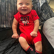 Rickey is registered to the contest to win money with this photo: baby, smiling, red_clothing, sitting, bed, pillow, blanket, infant, happy, face, person, child, indoors, sock, cute, young, portrait, comfortable, cozy, nike