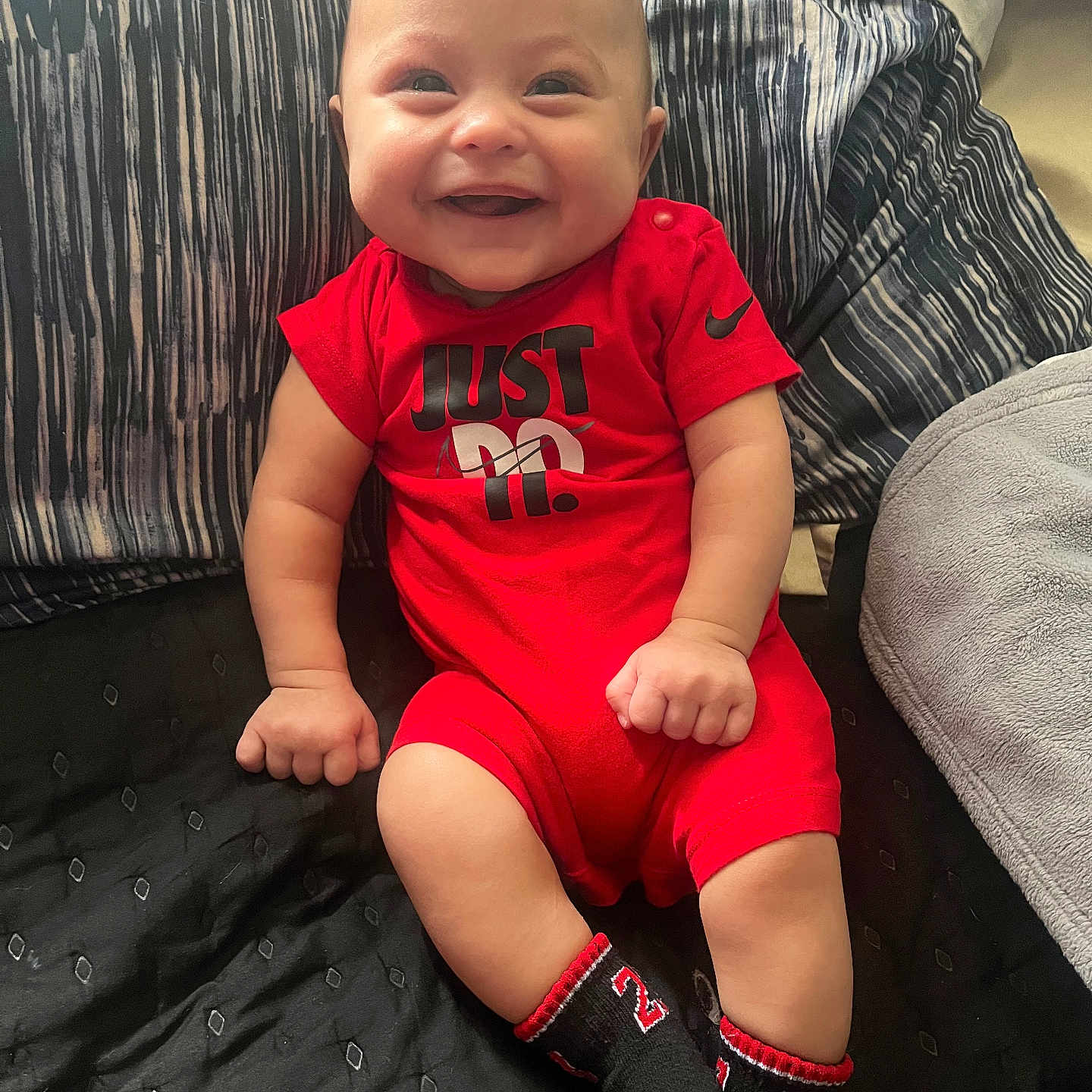 Rickey is registered to the contest to win money with this photo: baby, bed, blanket, child, comfortable, cozy, cute, face, happy, indoors, infant, nike, person, pillow, portrait, red_clothing, sitting, smiling, sock, young