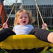 Stormi joined the competition — help win amazing prizes! child, swing, playground, outdoor, backyard, grass, fence, rope, happy, blonde_hair, white_shirt, black_pants, sunlight, fun, motion, person, swing_set, wooden_frame, smile, excited