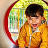 Joud Rayan a rejoint le concours — aidez-le/la à gagner de superbes lots ! child, toddler, yellow_jacket, smiling, playground, tunnel, outdoor, daylight, happy, crawling, face, hair, hands, fence, trees, park, play, casual_clothing, fun, cute