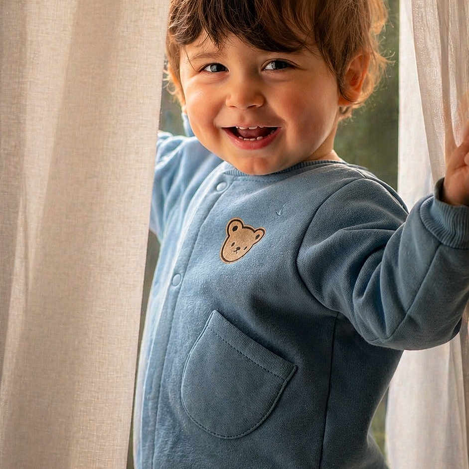 Joud Rayan a rejoint le concours — aidez-le/la à gagner de superbes lots ! child, toddler, smiling, curtains, blue_jacket, bear_patch, brown_pants, indoor, natural_light, window, happy, cute, portrait, clothing, young_child, person, face, hair, standing, playful