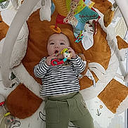 Lilio a rejoint le concours — aidez-le/la à gagner de superbes lots ! baby, infant, toy, playmat, plush_toy, teething_ring, striped_shirt, green_pants, hand, giraffe_toy, soft_toy, floor_mat, child, cute, indoor, playtime, colorful, person, lying_down, curious